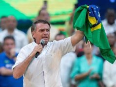 Jair Bolsonaro com camisa social branca, falando em microfone e segurando bandeira do Brasil
